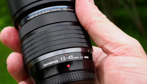 Hand holding an Olympus 12-45mm f3.5-5.6 telephoto zoom lens against a blurred green background.
