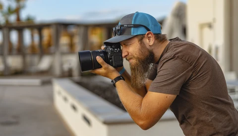 Bearded photographer in blue cap and brown shirt shooting with a DSLR camera on an urban rooftop.