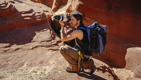 Adventure photographer crouching in desert canyon while photographing with telephoto lens and backpack.