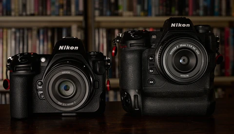 Two black Nikon DSLR cameras side by side, with the smaller model on the left and larger model on the right, displayed against a blurred bookshelf background.