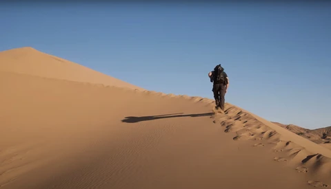 Hiker ascending a steep sand dune under clear blue sky, casting a long shadow on the golden slope.