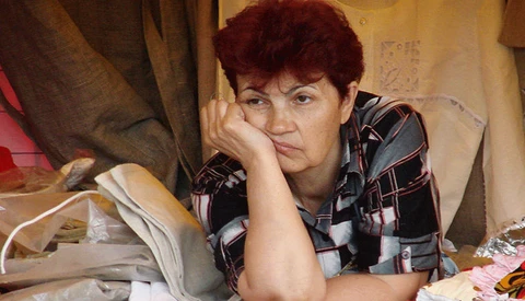 Woman with short reddish hair resting her head on her hand, sitting among fabric and materials in an indoor workspace.