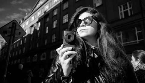 Woman in sunglasses holding a vintage camera on an urban street with period buildings behind her.