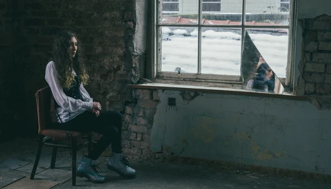 Person seated on a wooden chair in an industrial room, gazing toward a window overlooking a frozen landscape.