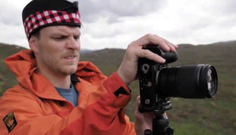 Photographer in orange jacket and red plaid headband holding a DSLR camera with telephoto lens in an open moorland landscape.