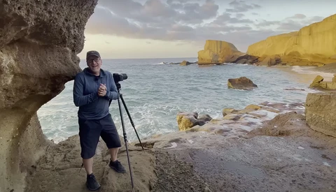 Photographer with tripod-mounted camera shooting coastal rock formations during golden hour.