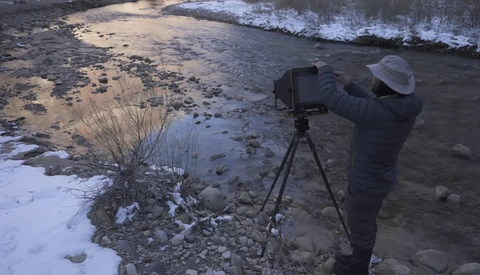 Photographer operating a camera on tripod at the edge of a rocky shoreline with incoming waves.
