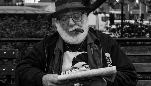 Older man with white beard wearing glasses and a hat, smoking a cigar while holding a newspaper on a park bench.