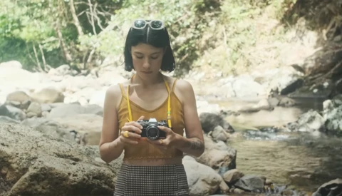 Woman in yellow top holding a vintage camera by a rocky stream.