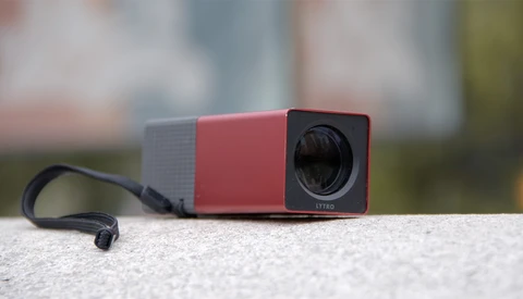 Compact red and gray projector with lens visible, sitting on concrete surface with blurred background.