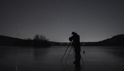 Silhouetted photographer on frozen lake at night with telescope on tripod pointed at starry sky.