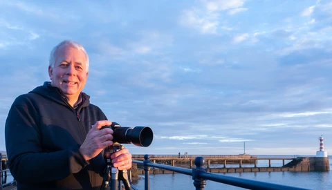 Photographer holding a telephoto lens camera on a tripod overlooking a harbor at golden hour.
