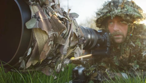 Soldier in ghillie suit holding rifle with large telephoto lens attachment, lying in grass.