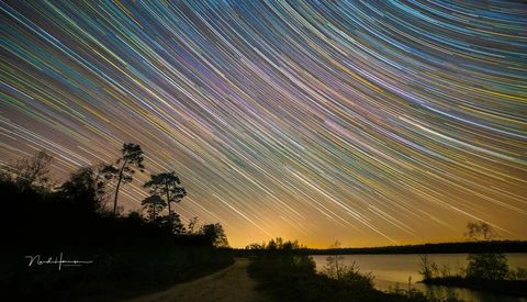 Star trail arcs sweep across the night sky above a dark lakeside landscape with silhouetted trees.