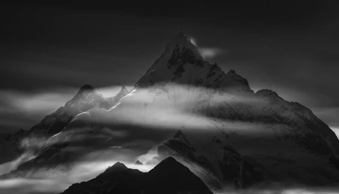 Dramatic black and white mountain landscape with mist swirling around jagged peaks beneath dark clouds.