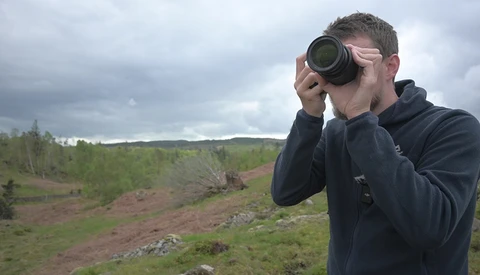 Photographer holding binoculars to his eyes while standing in a rolling landscape.