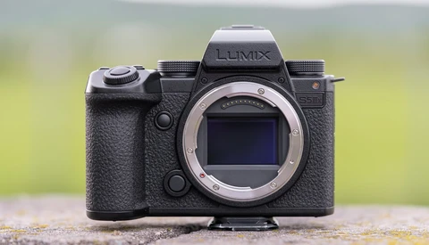 Panasonic Lumix mirrorless camera body with lens mount visible, positioned on a textured surface with blurred green background.