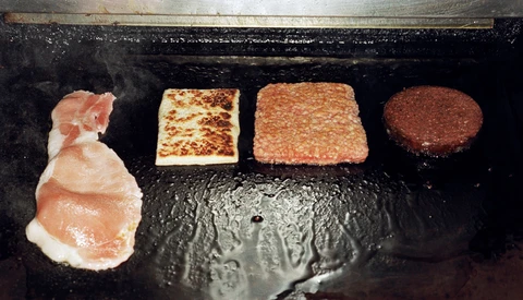 Four food items at different cooking stages on a dark griddle: raw prosciutto, lightly cooked patty, medium-cooked patty, and fully cooked dark patty.