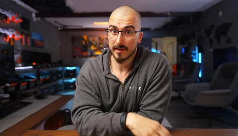 Man with glasses wearing a gray hoodie in a gaming setup studio with RGB lighting and multiple monitors.