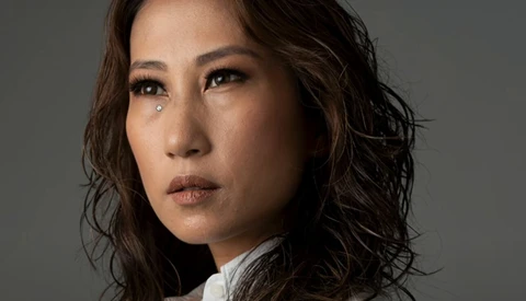 Portrait of a woman with wavy dark hair wearing a white shirt, gazing to the side against a neutral gray background.