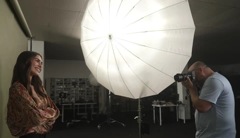 Photographer using a camera on tripod while model stands under large umbrella softbox in studio setting.