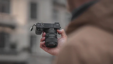 Photographer holding a rangefinder camera with attached lens, shot outdoors with blurred building backdrop.