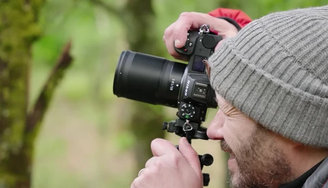 Photographer holding a DSLR camera with telephoto lens while looking through viewfinder outdoors.