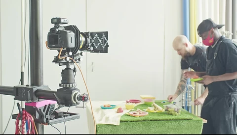 Professional video camera mounted on tripod filming two people preparing food at a table with green surface.