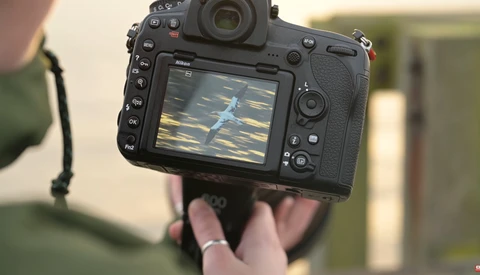 Photographer holding a Nikon DSLR camera displaying an aerial shot of a landscape on the rear LCD screen.