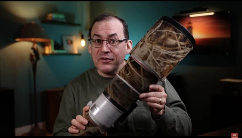 Man with glasses holding a camera lens and its protective case in a studio setting.