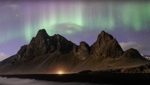 Jagged volcanic mountains silhouetted against a green and purple aurora borealis display over dark water.