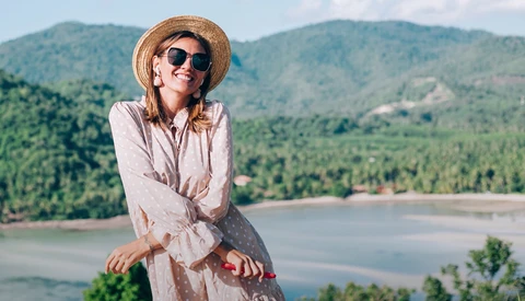 Woman in light shirt and straw hat smiling at camera overlooking forested mountains and river valley.