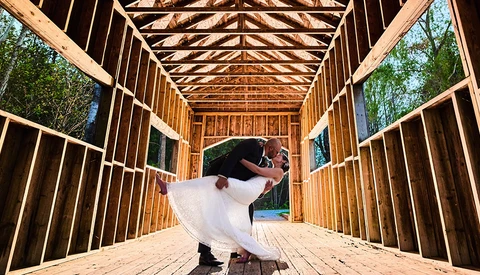 Bride in white dress twirling inside wooden barn structure with open sides and geometric roof framing.
