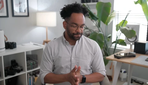 Man in light gray button-up shirt gesturing while speaking in a bright home office with plants and shelving visible behind him.