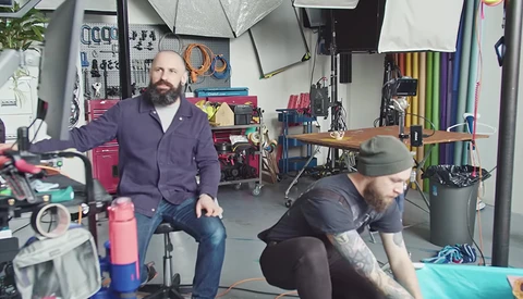 Two people in a workshop setting surrounded by photography and creative equipment on shelves and walls.