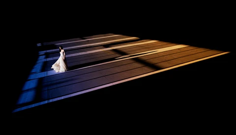 Bride in white dress walking alone on dark wooden floor with dramatic blue and gold directional lighting.