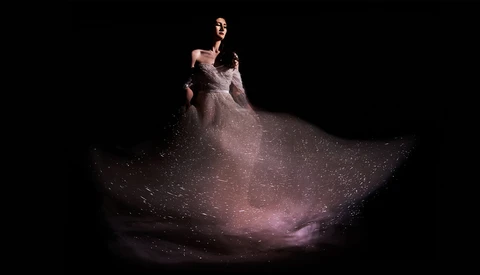 Woman in flowing, sparkle-embellished gown poses against dark background with dramatic lighting.