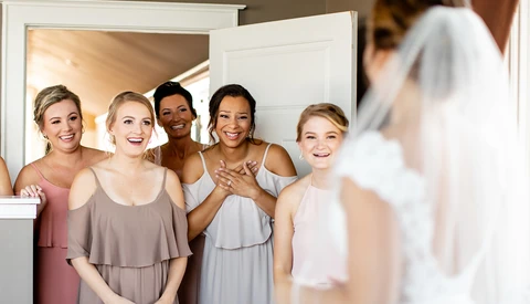 Bride in white dress viewed from behind as bridesmaids in pastel dresses smile at camera.