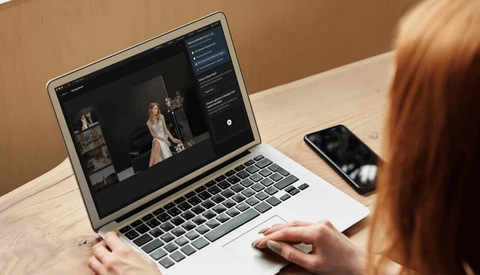 Photographer editing portrait images on laptop with smartphone nearby at wooden desk.