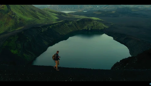 Hiker in red jacket standing on dark volcanic terrain overlooking a turquoise crater lake surrounded by green mountains.