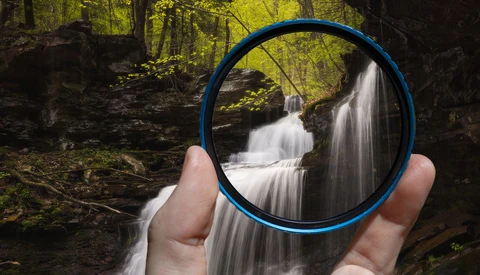 Hand holding blue circular filter frame against cascading forest waterfall.