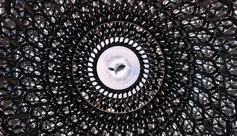Looking up through a geometric lattice structure at a circular opening framing the sky and birds in flight.