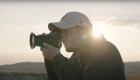 Videographer holding a cinema camera against backlit golden hour sky with hills in distance.