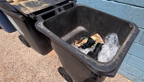Two black wheeled trash bins against a blue wall, one partially open with crumpled plastic and cardboard inside.