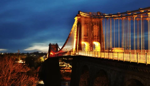 Suspension bridge illuminated in golden light at dusk against a dramatic blue sky.