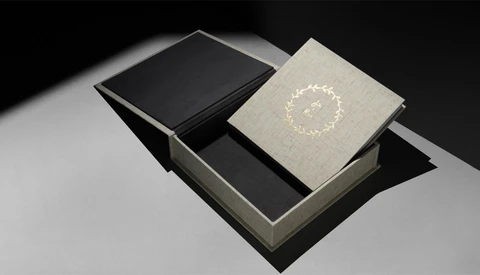 Luxury book packaging with a cream linen-covered volume featuring a decorative wreath design, displayed with a black book and gray box.