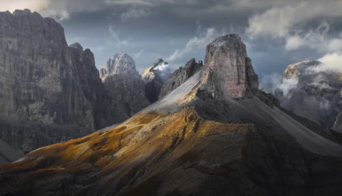 Dramatic mountain landscape with layered peaks and golden-lit slopes beneath stormy clouds.