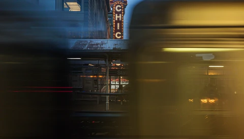 Motion-blurred subway train passing in front of historic theater marquee at dusk.