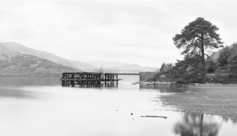 Wooden boathouse structure reflected in still water with forested shoreline and mountains beyond.