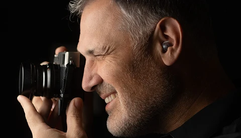 Male photographer holding binoculars-style camera to eyes against black background.